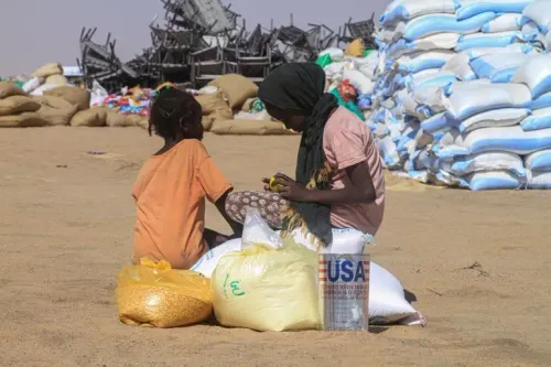 امرأتان جالستان على أكياس من المساعدات الغذائية في منطقة صحراوية، وسط أكياس أخرى، تعكسان أزمة إنسانية متزايدة في السودان.