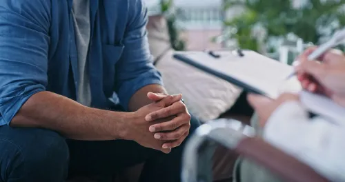 رجل يجلس في جلسة استشارية نفسية، مع التركيز على يديه المتشابكتين، بينما يكتب المُعالج ملاحظات على دفتر.