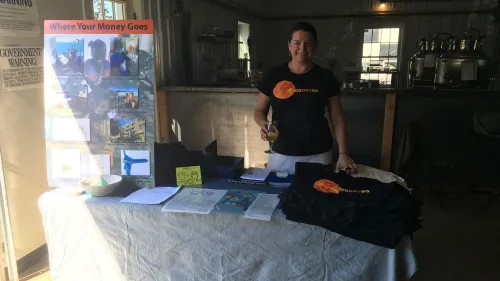 امرأة تحمل كأسًا، تقف أمام طاولة مغطاة بقماش، حيث تعرض مواد توعوية حول الرعاية الصحية الإنجابية وحقوق الإجهاض.