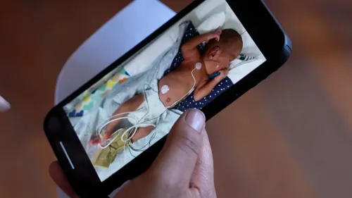 صورة تُظهر والد يحمل هاتفًا يظهر فيه توأم حديث الولادة في وحدة العناية المركزة، مع أسلاك طبية على جسمه.