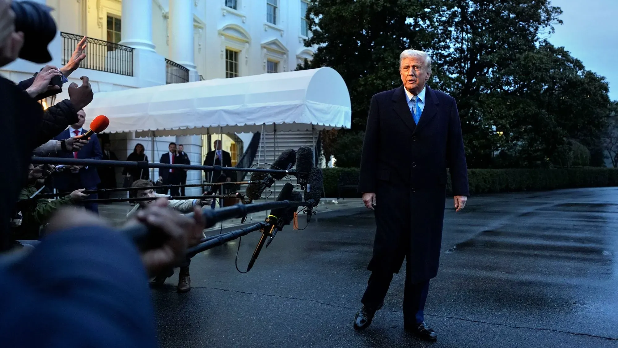 رئيس الولايات المتحدة السابق دونالد ترامب يتقدم نحو الصحفيين خارج البيت الأبيض، محاطاً بكاميرات وميكروفونات، في أجواء غائمة.