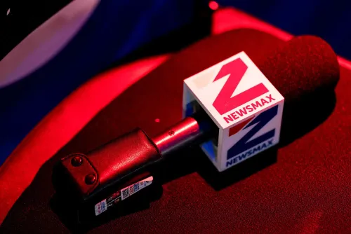 ميكروفون يحمل شعار نيوزماكس، مع إضاءة حمراء خلفية، يعكس التركيز على القناة في سياق تسوياتها القانونية المتعلقة بالانتخابات.