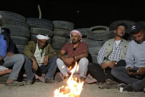 مجموعة من رجال قرية المفقرة يجلسون حول نار مشتعلة في الليل، محاطين بإطارات قديمة، يتبادلون الحديث أثناء حراستهم للقرية.