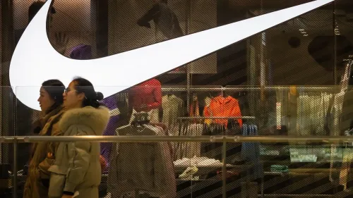 امرأتان تسيران بجانب واجهة متجر نايكي، حيث يظهر شعار الشركة الكبير والملابس الرياضية المعروضة.