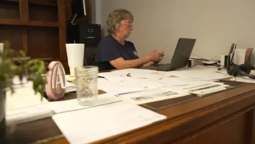 امرأة مسنّة تجلس أمام مكتب مليء بالملفات والأوراق، تستخدم الحاسوب المحمول وتفكر في تأثيرات إعصار هيلين على مجتمعها.