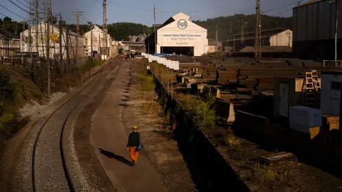 مشهد لمصنع يو إس ستيل، يظهر شخصًا يسير في ممر بجوار المباني الصناعية، مع لافتة تحمل اسم الشركة، مما يعكس التحديات التي تواجهها الشركة.