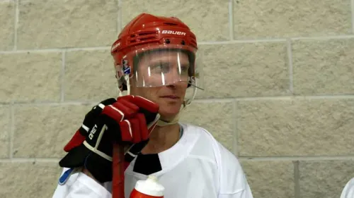 لاعب هوكي يرتدي خوذة حمراء وقميص أبيض، يضع يده على ذقنه في موقف تأملي، مع خلفية جدار رمادي.