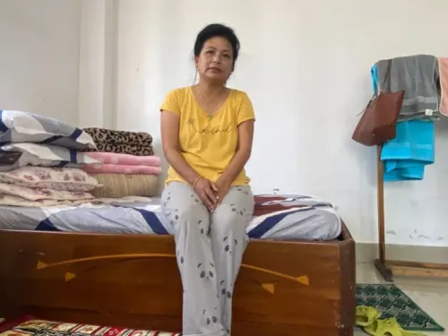 امرأة تجلس على سرير في غرفة بسيطة، محاطة بوسائد وأغطية، تعكس آثار الفيضانات في سيكيم.