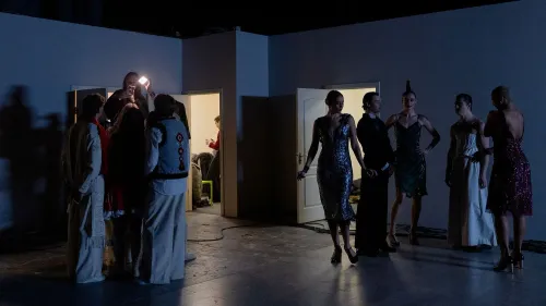 مجموعة من الأشخاص يرتدون ملابس أنيقة في كواليس عرض أزياء، مع إضاءة خافتة وظلال واضحة، مما يعكس أجواء التحضير للعرض.