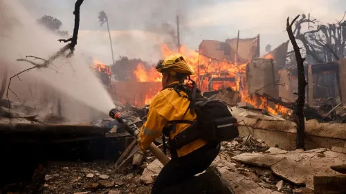 رجل إطفاء يستخدم خرطومًا لمكافحة حرائق الغابات في كاليفورنيا، بينما تتصاعد ألسنة اللهب من المباني المدمرة خلفه.