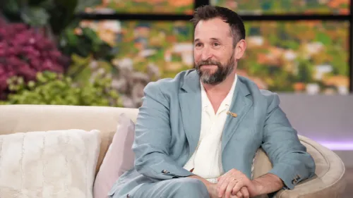 نواه وايل يجلس مبتسمًا على الأريكة في برنامج "ذا جينيفر هدسون شو"، مع خلفية ملونة من الزهور والنباتات.