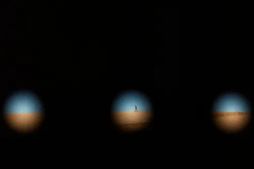 رجل يسير في صحراء تحت سماء زرقاء، يظهر من خلال ثلاث فتحات داكنة، مما يرمز إلى قضايا الهجرة والتحديات السياسية.