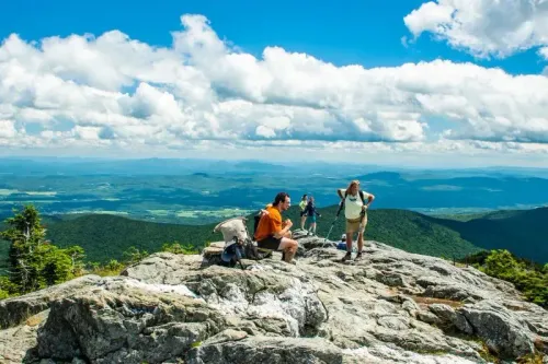مجموعة من المتنزهين يستمتعون بالمناظر الطبيعية الخلابة من قمة جبل في منطقة نورث كونتري، نيويورك، تحت سماء زرقاء مع غيوم بيضاء.