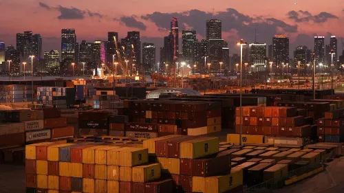 صورة لميناء مزدحم في مدينة كبيرة، حيث تكدست حاويات الشحن تحت سماء غائمة، مما يبرز تأثير التعريفات الجمركية على التجارة.