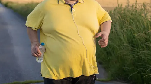 رجل بدين يرتدي قميصًا أصفر، يسير في الهواء الطلق ويمسك بزجاجة ماء، مما يعكس قضايا التثدي والوزن الزائد.