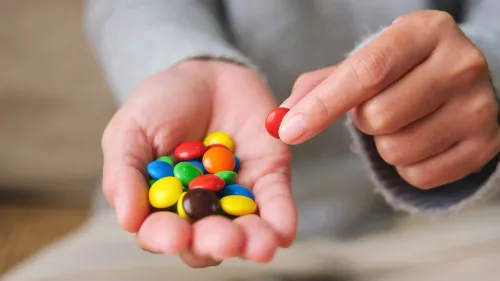 يد تحمل حلوى ملونة متنوعة، بما في ذلك الأحمر والأصفر والأخضر، في سياق مناقشة حول المخاطر الصحية للملونات الغذائية.