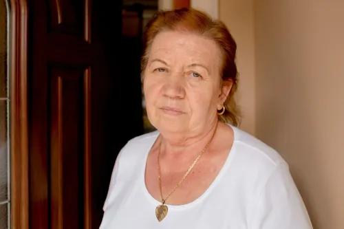 امرأة مسنّة تحمل تعبيرًا جادًا، تقف أمام باب منزلها، تعكس مشاعر الخوف والقلق بعد حادثة إطلاق نار في مكان عملها.