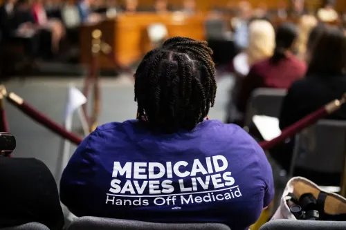امرأة ترتدي قميصًا أرجوانيًا يحمل عبارة "Medicaid Saves Lives" خلال جلسة استماع، تعبر عن دعمها لبرنامج Medicaid.