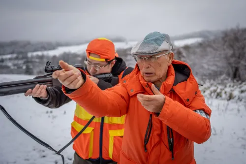 رجل مسن يرتدي سترة برتقالية زاهية، يشير بيده بينما يتدرب على صيد الدببة في منطقة جبلية مغطاة بالثلوج في هوكايدو، اليابان.