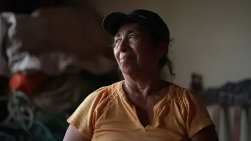 امرأة مسنّة مبتسمة ترتدي قميصًا برتقاليًا، تجلس في مكان مظلم محاط بأدوات الصيد، تعكس ملامحها عمق التجارب الحياتية.