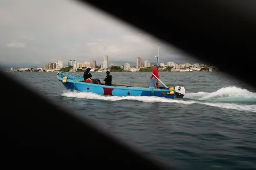 قارب صيد ملون يحمل ثلاثة رجال يرتدون ملابس سوداء، يبحر في المياه بالقرب من مدينة ساحلية، مع خلفية من المباني العالية.