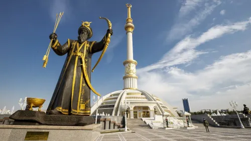 تمثال ذهبي يمثل شخصية تاريخية في عشق آباد، بجوار معلم معماري حديث، تحت سماء زرقاء. يعكس التباين بين التراث والحداثة في تركمانستان.