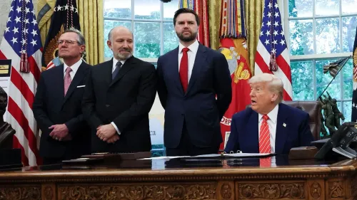 الرئيس ترامب يجلس خلف مكتبه في المكتب البيضاوي، محاطًا بمساعدين، أثناء توقيع اتفاق تجاري مع المملكة المتحدة، مما يعكس جهود الإدارة لتعزيز العلاقات التجارية.