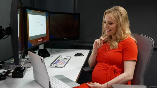 امرأة حامل ترتدي فستانًا أحمر، تجلس أمام جهاز كمبيوتر محمول، مبتسمة وتبدو مشغولة بالتفاعل مع الشاشة.