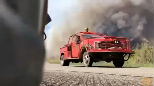 سيارة إطفاء حمراء تسير على طريق، بينما تتصاعد سحب الدخان الكثيف من حريق غابات في نيوجيرسي، مما يهدد المباني ويؤدي إلى إجلاء السكان.