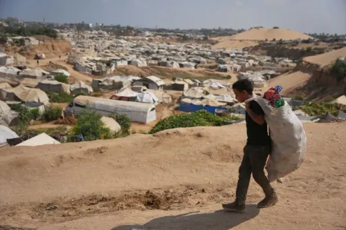 شاب فلسطيني يحمل حقيبة كبيرة على ظهره أثناء سيره في مخيم للنازحين في قطاع غزة، يعكس تداعيات النزاع والتهجير المستمر.