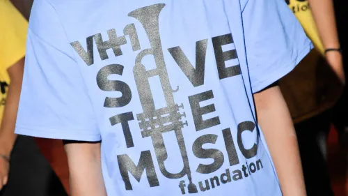 تيشيرت أزرق يحمل شعار "Save the Music" مع تصميم يبرز أداة موسيقية، يرمز لدعم التعليم الموسيقي في المدارس.