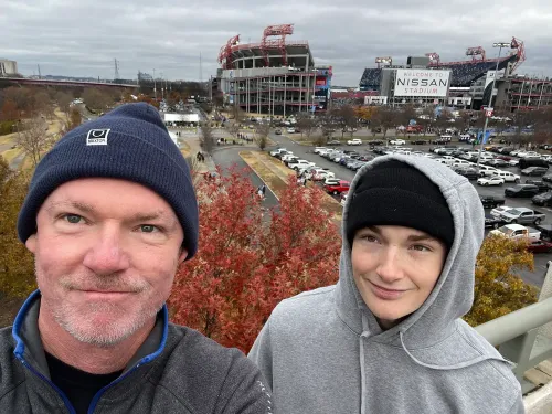 صورة لوالد ونجله في ملعب نيسان، مع خلفية لمبنى الملعب وأشجار ملونة، تعكس لحظة عائلية وسط التحديات النفسية.