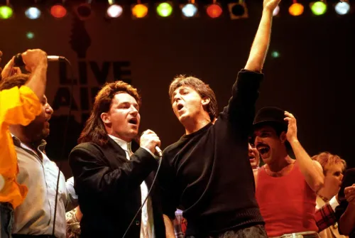 مكارتني يغني "Let It Be" في حفل Live Aid، محاطًا بفنانين مثل بونو وفريدي ميركوري، وسط أجواء حماسية.