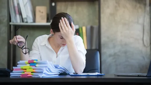 امرأة ترتدي قميصًا أبيض وتجلس على مكتب، تبدو مرهقة وتفكر بقلق وسط أوراق مرتبة بشكل عشوائي، تعكس الضغط الناتج عن الكمالية.