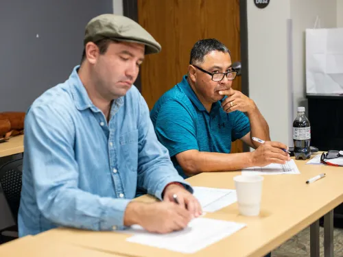 رجلان يجلسان في غرفة دراسية، يكتبان ملاحظات خلال جلسة شعرية مخصصة للمصابين بالخرف وأسرهم، مع التركيز على أهمية التعبير عن الذات.