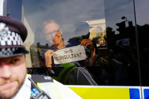رجل مبتسم يحمل لافتة مكتوب عليها "استشاري NHS" داخل سيارة شرطة، بينما يظهر ضابط شرطة في المقدمة خلال مظاهرة في لندن.