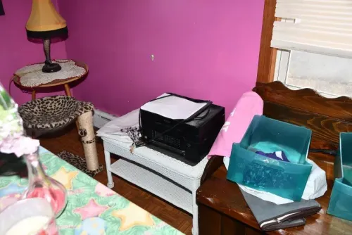 غرفة نوم ذات حائط وردي، تحتوي على طابعة وأشياء عشوائية، تعكس حالة الإهمال داخل منزل المحتجز.