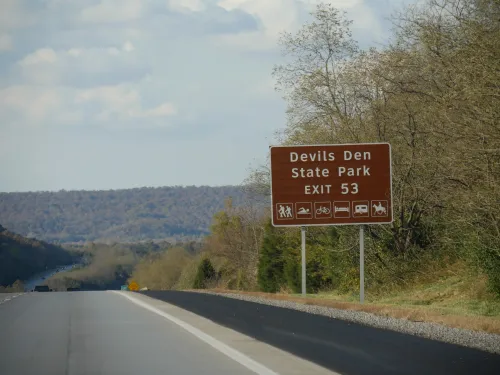 علامة طريق تشير إلى متنزه ديفلز دن ستيت بارك في أركنساس، مع خلفية طبيعية جبلية، حيث وقع حادث مأسوي مؤخرًا.