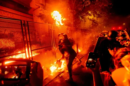 محتجون يشعلون النار في سياج خلال مظاهرة في بورتلاند، مع وجود أشخاص يحملون هواتفهم لتوثيق الحدث وسط أجواء مشحونة.