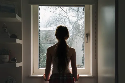 فتاة صغيرة تقف أمام نافذة، تنظر إلى الخارج في ضوء النهار، تعكس مشاعر العزلة والقلق المرتبطة بزيادة حالات إيذاء النفس بين الأطفال.