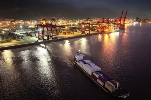 صورة لميناء بحري ليلاً، حيث تظهر سفينة شحن تحمل حاويات، مع وجود رافعات ومخازن مضاءة في الخلفية. تعكس الصورة تأثير الرسوم الجمركية على التجارة.