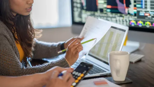 امرأة تستخدم حاسوبًا محمولًا أثناء مراجعة مستندات مالية، مع شاشة تعرض بيانات سوقية في الخلفية، تعكس أهمية الاستثمار في الأسهم الخاصة.