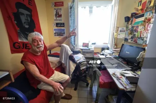 جورج إبراهيم عبد الله، المناضل اللبناني، يجلس في غرفة مليئة بالكتب والصور، مشيرًا بيده نحو النافذة، مع وجود علم فلسطيني خلفه.