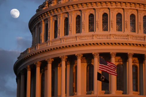 قبة مبنى الكابيتول الأمريكي تضيء بأشعة الشمس الغاربة، مع علم الولايات المتحدة يرفرف، والقمر في السماء، مما يرمز إلى أهمية السياسة في الانتخابات القادمة.