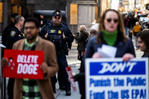 محتجون يحملون لافتات تدعو إلى حذف "دوج" في وسط المدينة، بينما يتواجد ضابط شرطة في الخلفية، مما يعكس التوترات حول سياسات الحكومة الفيدرالية.