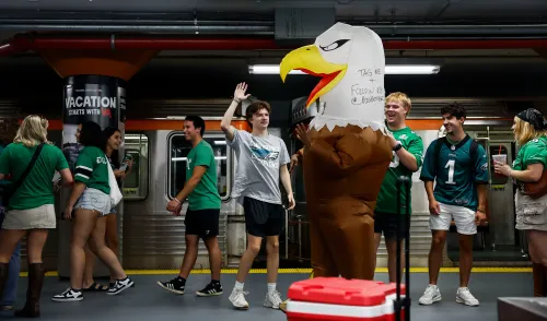 مشجعو فريق فيلادلفيا إيجلز يرتدون قمصان الفريق في محطة قطار، مع شخصية تمثل النسر، استعدادًا لمباراة.