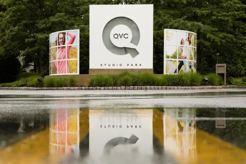 علامة QVC في حديقة الاستوديو، محاطة بصور ترويجية لموظفين، تعكس تاريخ الشركة في التسوق المباشر وتحدياتها الحالية.