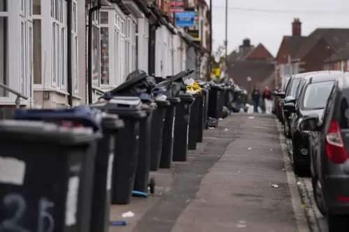 تظهر الصورة صفًا من حاويات القمامة الممتلئة على جانب طريق في برمنغهام، مما يعكس أزمة تراكم النفايات في المدينة.