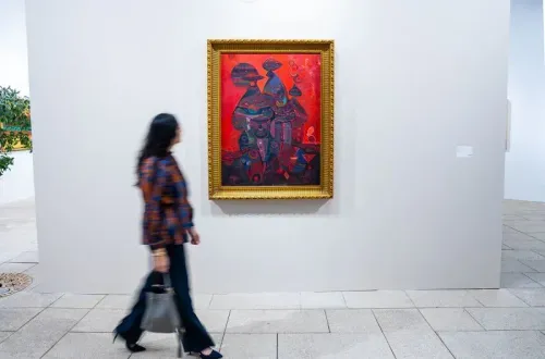 امرأة تسير بجانب لوحة فنية ملونة في إطار ذهبي، تعرض ألوانًا دافئة ونقوشًا معقدة، في معرض فني بالمملكة العربية السعودية.