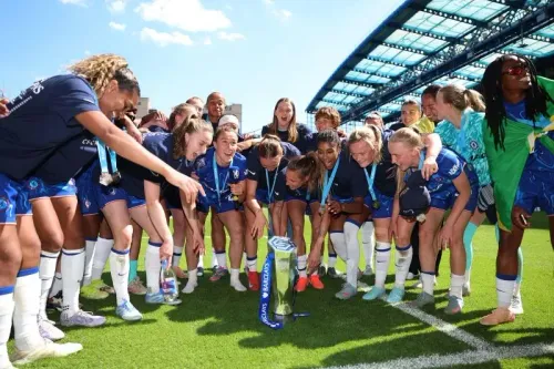 فريق تشيلسي للسيدات يحتفل بفوزهم بالبطولة، مع عرض الكأس في وسط الصورة، بينما يبتسم اللاعبون ويعبرون عن فرحتهم.